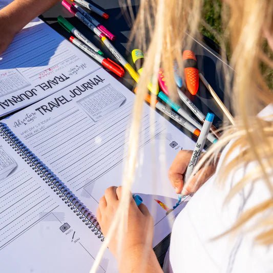 Young girl filling in her Adventure Awaits Kids Travel Journal outdoors using colourful Sharpie markers, shown open to the journal entry pages